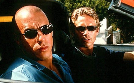 Vin Diesel e Paul Walker dentro de um carro, de óculos escuros, olhando para o lado