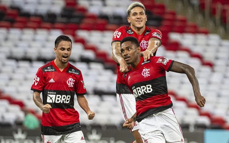 Michael celebra com punhos cerrados enquanto Pedro pula em cima de Vitinho para comemorar gol do Flamengo; todos estão com o uniforme de jogo principal vermelho e preto
