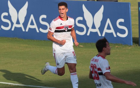 Vitor Bueno com uniforme branco do São Paulo corre com punhos cerrados em comemoração de gol e outro jogador desfocado aparece na parte de baixo
