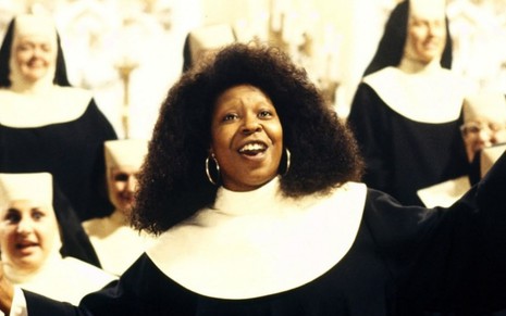 Em frente a algumas freiras, a atriz Whoopi Goldberg canta e usa roupa de freira em cena do filme Mudança de Hábito