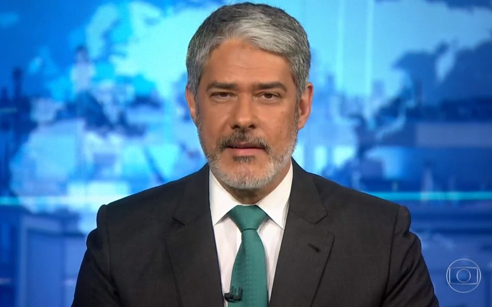 William Bonner com terno escuro, camisa branca e gravata verde na bancada do Jornal Nacional