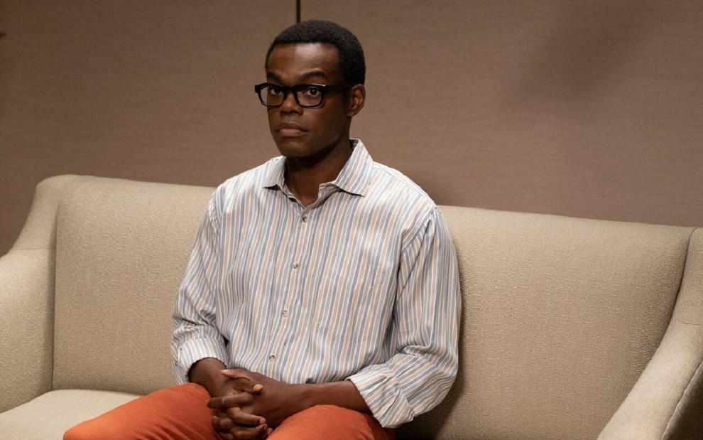 Chidi Anagonye, personagem de William Jackson Harper, está sentado em um sofá em uma cena do seriado The Good Place. Ele é negro, está usando óculos de armação preta e uma camisa listrada