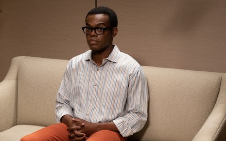 Chidi Anagonye, personagem de William Jackson Harper, está sentado em um sofá em uma cena do seriado The Good Place. Ele é negro, está usando óculos de armação preta e uma camisa listrada