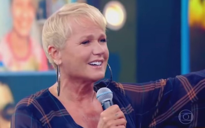 Xuxa Meneghel em participação no Caldeirão do Huck, de camisa xadrez, com o microfone na mão direita