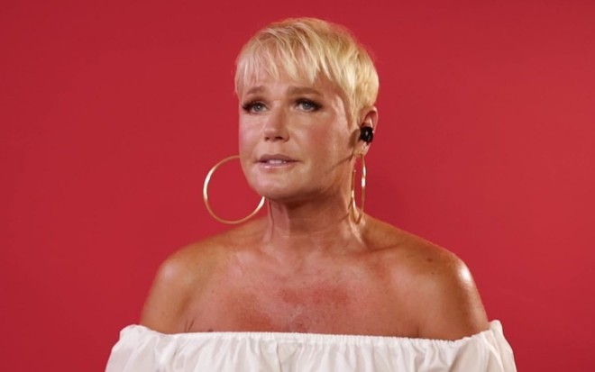 Xuxa com dois brincos de argola e uma blusa branca ombro a ombro, em fundo vermelho