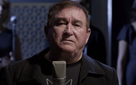 Em pé, Amado Batista veste um casaco e uma camiseta preta e está na frente de um microfone de um estúdio de gravação 