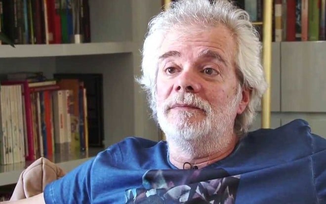 O autor Carlos Lombardi, de camiseta azul estampada, dá entrevista em uma poltrona de sua casa