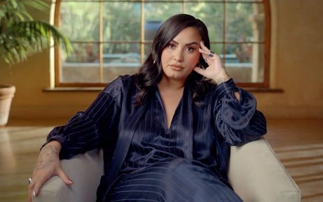 Com uma roupa de seda, Demi Lovato está sentada no sofá de sua casa, com o rosto apoiado na mão esquerda, em cena da série documental Dancing With the Devil