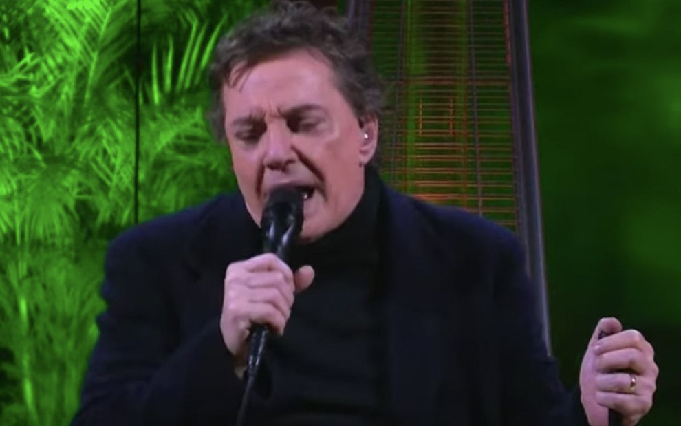 O cantor Fábio Jr. em live do dia dos namorados: música canta com um microfone preto e usa casaco preto