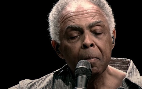 Gilberto Gil canta com o olhos direcionados para o chão e usa camisa cinza 