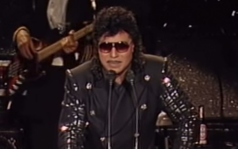 Little Richard com uma roupa preta brilhosa de óculos escuros em frente a dois microfones em uma tribuna