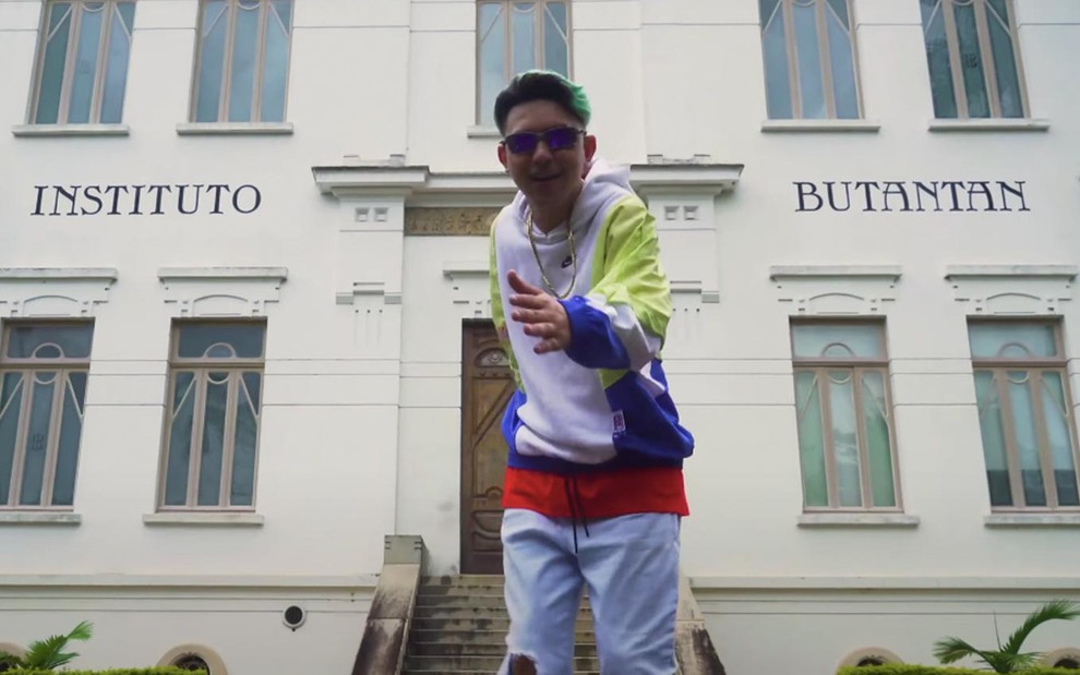 MC Fioti em clipe do Remix Vacina Bum Bum Tan Tan: funkeiro está com óculos azul espelhado, cabelo colorido, moletom branco e verde e usa corrente de ouro