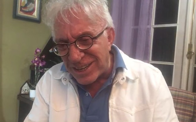 Com camisa azul e jaqueta branca, Moacyr Franco sorri durante live no YouTube