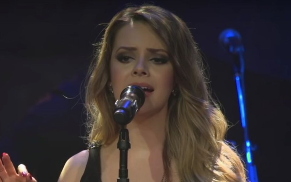 Em pé em um palco, a cantora Sandy canta diante de um microfone instalado em um pedestal