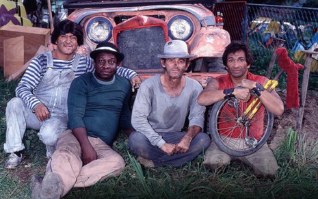 Zacarias, Mussum, Renato Aragão e Dedé Santana sujos e suados, sentados lado a lado, em frente a um carro