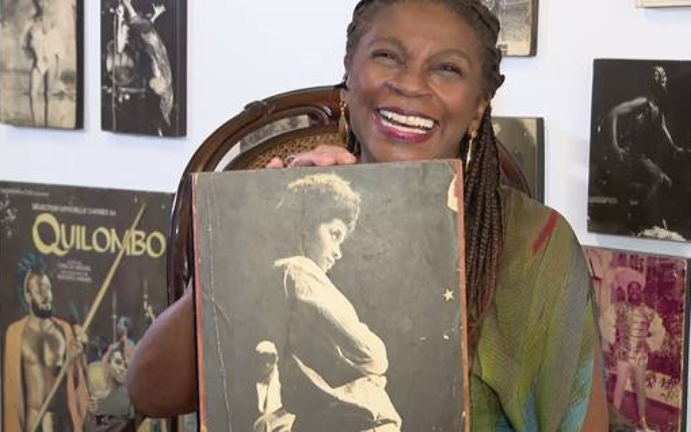 Zezé Motta sentada, sorrindo, segurando um quadro com uma pessoa negra, atrás dela há uma parede branca com outros de quadros com pessoas negras