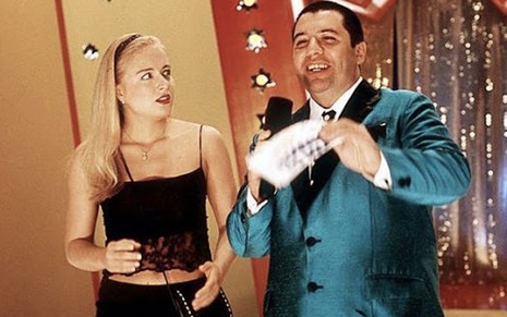 A apresentadora Angélica olha para a direita como a Angel e observa Bussunda com um terno de cetim azul, ele segura uma ficha com a mão direita e um microfone com a esquerda , em cena de Zoando na TV