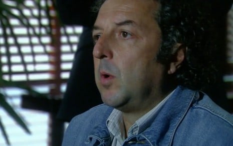 O ator Luiz Ramalho como Baiano em A Favorita; ele está sentado, de lado, com a boca semiaberta ernquanto conversa durante um depoimento da cena