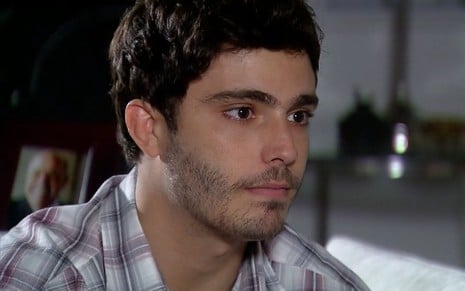O ator Thiago Rodrigues como Cassiano em A Favorita; ele está sentado, olhando para o lado com cara séria