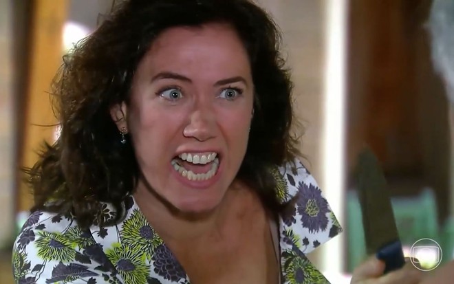 A atriz Lilia Cabral como Catarina em A Favorita; ela está de lado, olhando para frente com uma faca apontada e a boca aberta enquanto briga