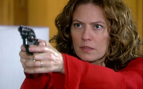 A atriz Patricia Pillar como Flora em A Favorita; ela está segurando uma arma apontada em sua frente enquanto olha fixamente com cara séria