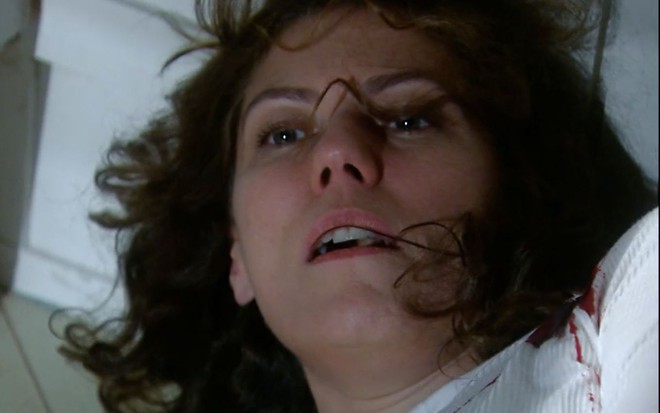 A atriz Patricia Pillar como Flora em A Favorita; ela está deitada no chão, olhando para cima com cara de dor e manchada de sangue após levar um tiro no peito