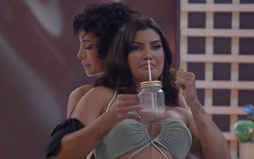 Marina usa um vestido verde, segura um copo transparente e está de costas para Aline
