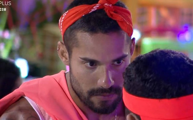 Bil olha para Rico, veste regata laranja e lenço laranja na cabeça