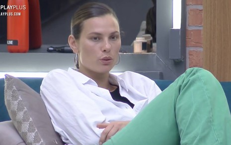 Dayane Mello veste camiseta branca e calça verde; ela está sentada em um sofá azul