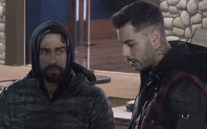 Erasmo está sentado ao lado de Gui, ele veste uma jaqueta preta e olha para o lado; Gui olha para baixo e veste jaqueta preta