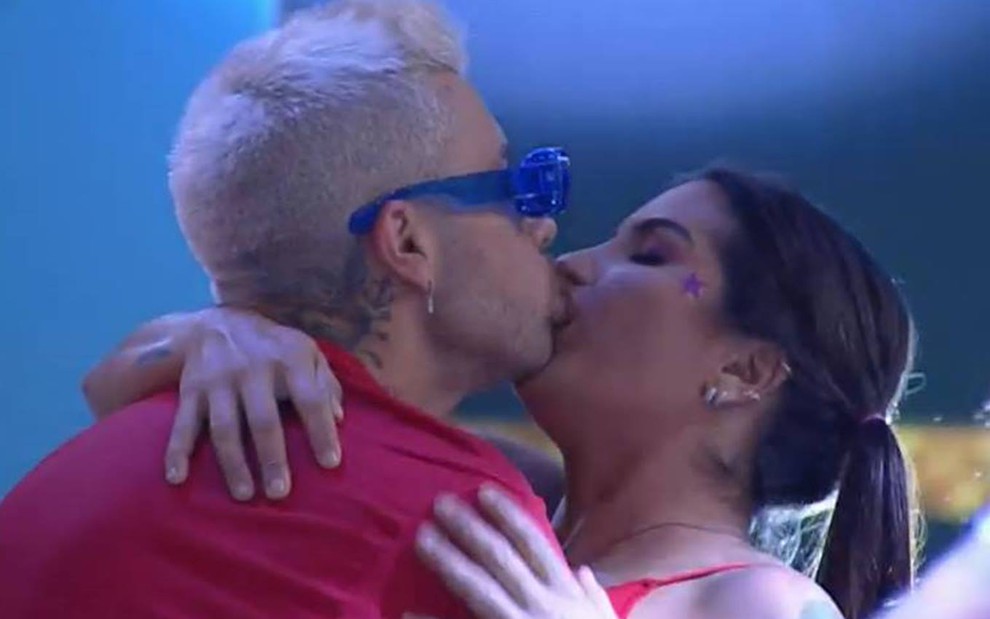Gui está beijando Marina; ele veste camiseta vermelha e usa óculos azul; Marina veste regata vermelha e está com o cabelo preso