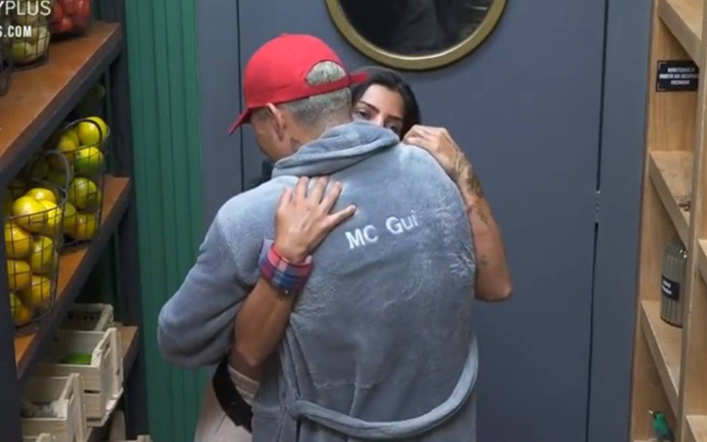 Marina está abraçada com MC Gui; ele está de costas, usa um boné vermelho e veste um roupão cinza