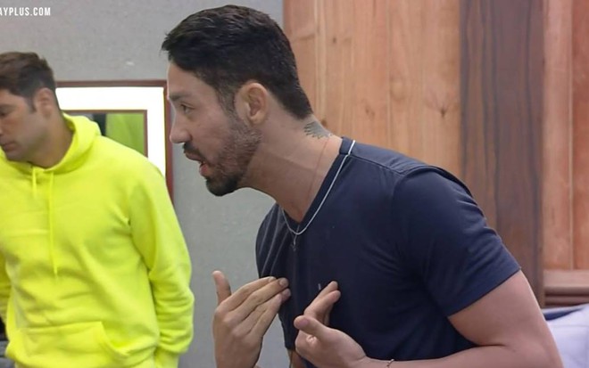Rico olha para o lado, veste camiseta azul e está com as duas mãos no peito