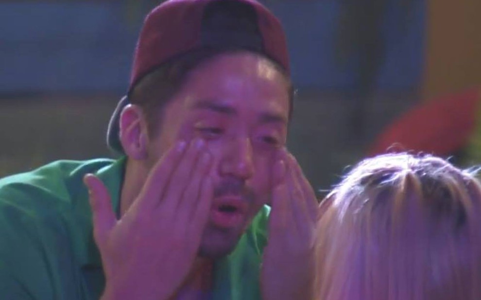 Rico está com as mãos no rosto, chora e veste camiseta verde
