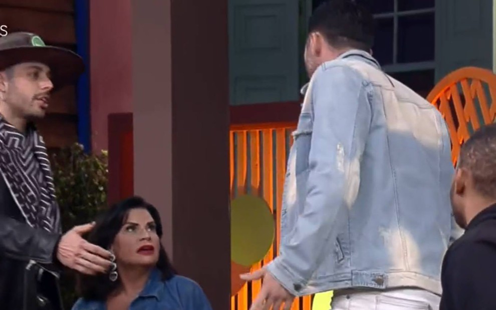 Valentina está sentada, ela olha para Rico; ele está em pé, olha para a peoa e veste jaqueta jeans