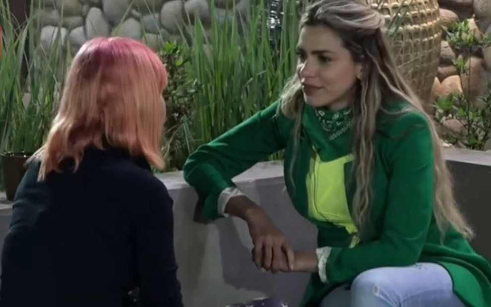 Valentina está sentada na frente de Erika, veste blusa preta e está com o cabelo solto; Erika olha para Valentina, veste blusa verde e camiseta verde