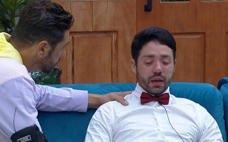 Rico está sentado, veste camiseta branca com gravata vermelha; Victor está ao lado de Rico e veste moletom colorido