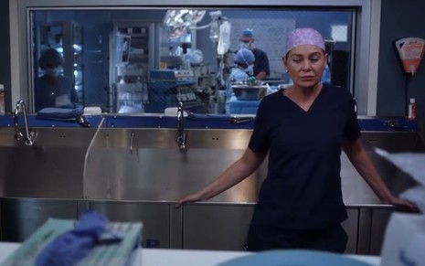 Ellen Pompeo está com roupa de cirurgião e apoiada na pia onde médicos lavam suas mãos, com expressão triste