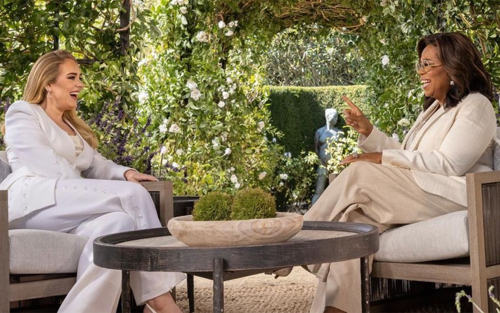 Cantora Adele e apresentadora Oprah Winfrey conversam sentadas em sofás de frente uma para outra