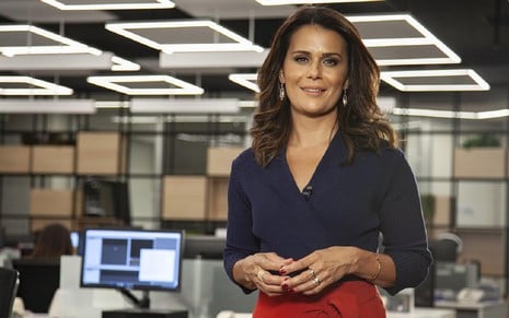 Adriana Araújo usa uma blusa azul com calça preta e sorri para a câmera. Ela está na redação da Record, onde trabalhou entre 2006 e 2021.