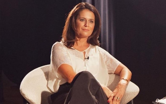Adriana Araújo de blusa branca e calça preta, sentada em uma poltrona branca, olhando fixamente para o lado