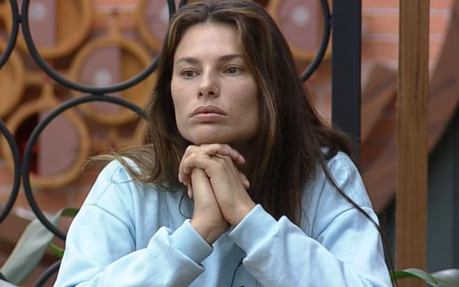 Dayane Mello em A Fazenda 13 com uma expressão reflexiva e a as duas mãos juntas apoiadas no queixo. Ela veste um moletom branco. No fundo é possível ver a decoração da sala do reality show.