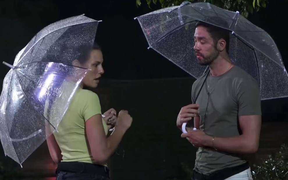 Imagem de Dayane Mello (à esq.) e Rico Melquiades usando guarda-chuva enquanto conversam em A Fazenda 13