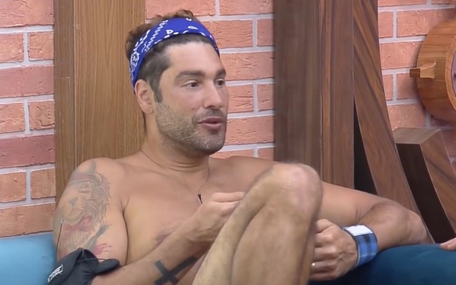 Imagem de Victor Pecoraro sem camisa e de bandana azul na sala de A Fazenda 13