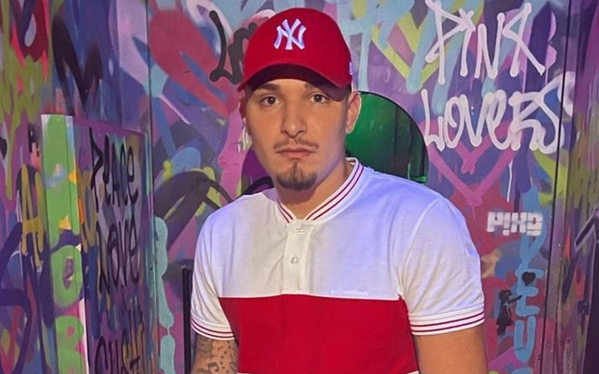 MC Gui está na frente de uma parede colorida, veste camiseta branca e com detalhe vermelho