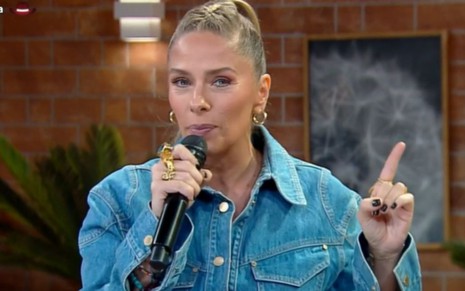 Adriane Galisteu segura um microfone preto, veste jaqueta jeans azul e está na frente de uma parede de tijolos