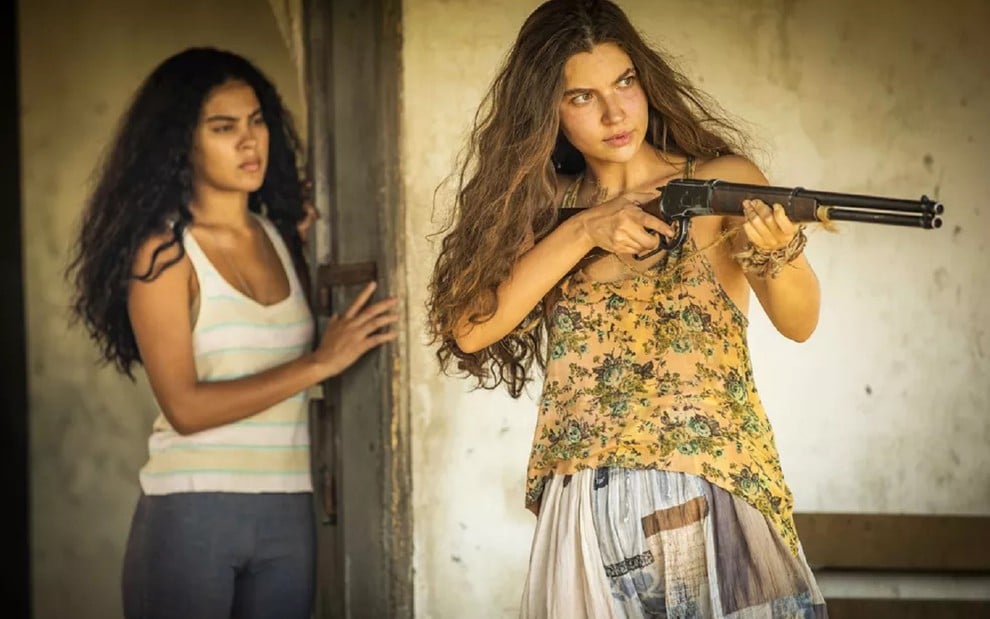 Alanis Guillen caracterizada como Juma Marruá, com uma blusa amarela uma arma em punho; Bella Campos como Muda ao fundo