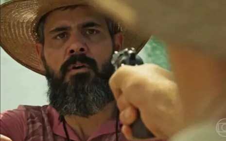 O ator Juliano Cazarré como Alcides em Pantanal; ele está de chapéu olhando com cara de assustado para uma arma, que está sendo apontada a sua frente