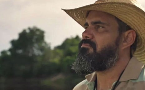 O ator Juliano Cazarré como Alcides em Pantanal; ele está de chapéu, virado para o lado olhando a paisagem com cara séria