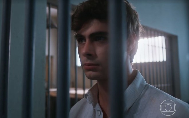 O ator Rafael Vitti como Davi em Além da Ilusão; ele está atrás das grades, olhando para frente com cara séria